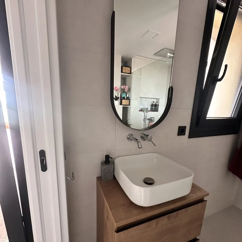 Baño reformado con lavabo sobre mueble de madera, grifería empotrada y espejo ovalado negro
