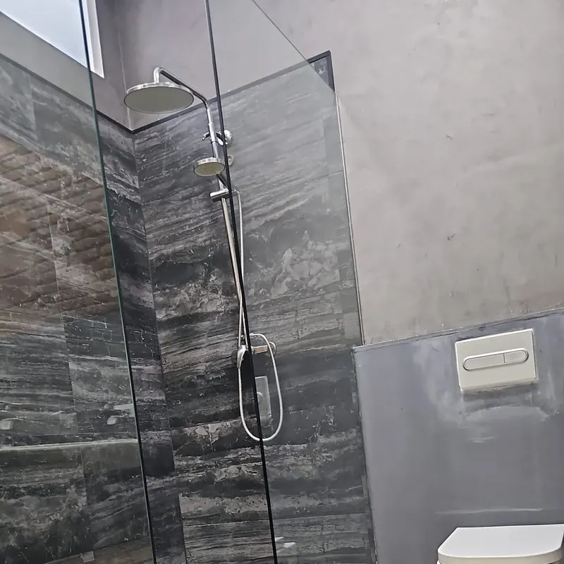 Ducha de obra a ras de suelo con porcelánico efecto mármol oscuro, mampara fija de cristal, columna de ducha e inodoro suspendido